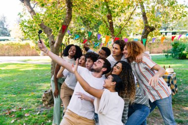 Bir grup neşeli genç öğrenci parkta parti sırasında akıllı telefon ve selfie çubuğuyla selfie çekiyorlar.