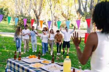 Bir grup neşeli çoklu etnik arkadaş bir parkta piknik masasına doğru yürüyor, kutlama yapıyor ve kollarını kaldırıyorlar.