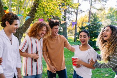 Neşeli bir grup genç arkadaş gülüyor, güneşli bir parkta eğleniyor, birlikteliği kutluyor ve neşeli anılar yaratıyorlar.