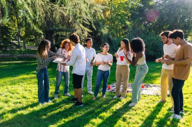 Neşeli çoklu etnik arkadaşlar abur cubur ve içecekleri paylaşıyor, parkta güneşli bir gün pikniğinin tadını çıkarıyor.