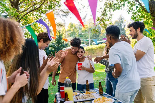 Bir grup neşeli çoklu etnik arkadaş açık hava partisinde yiyecek ve içeceklerle kucaklaşan genç bir çifti alkışlıyor ve kutluyorlar.