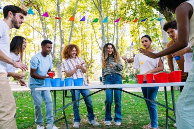 Bir grup neşeli, çoklu etnik arkadaş parkta bira pong oynuyor, açık havada güneşli bir günün tadını çıkarıyorlar.
