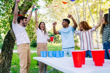 Beş çoklu etnik arkadaş silah kaldırıyor ve tezahürat yapıyor, dışarıda parkta bira pong oynayarak kazandıkları zaferi kutluyorlar.