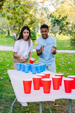 Genç bir adam ve kadın güneşli bir parkta canlı bir bira tenisi oynuyorlar. Kırmızı ve mavi bardaklara ustalıkla pin pon topu atıyorlar.
