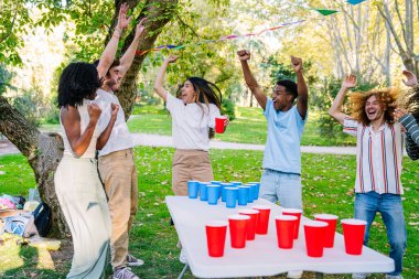 Neşeli çoklu etnik arkadaşlar bira pong zaferlerini kutluyor, neşeli bir park ortamında gülüp eğleniyorlar.