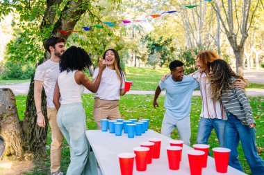 Neşeli çoklu etnik arkadaşlar parkta bira pong oynuyorlar güneşli bir günde eğleniyorlar ve eğleniyorlar.