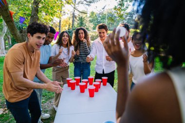 Neşeli çoklu etnik arkadaşlar neşe dolu bir bahçe partisinde açık havada bira tenisi oynuyorlar. Kahkaha ve heyecan dolu.