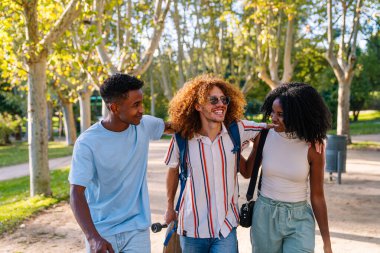 Üç neşeli çoklu etnik öğrenci parkta birlikte yürüyor, boş zamanlarının tadını çıkarıyor ve konuşuyorlar.