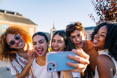 Bir grup neşeli çoklu etnik arkadaş şehirde akıllı telefonlarıyla selfie çekiyor, yaz tatilinin tadını çıkarıyorlar.