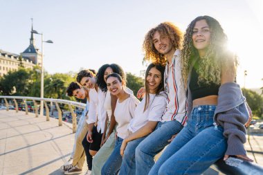 Mutlu çoklu etnik arkadaşlar parmaklıklara yaslanıyor, güneşli bir günün tadını çıkarıyor, kahkaha ve bağlantının görüntülerini yakalıyorlar.