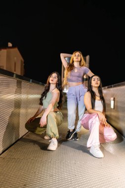 Üç genç kadın sokak dansçısı gece metal bir iskelede poz veriyor, kent tarzı ve dans ekibi arkadaşlıklarını sergiliyorlar.