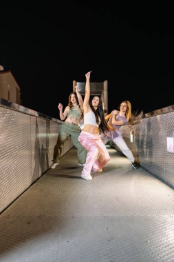 Metal bir köprüde sokak dansı becerilerini sergileyen bir grup genç kadın gece gökyüzünün altında enerjik bir gösteri yapıyorlar.