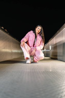 Gece çömelmiş metal bir köprüde poz veren sokak dansçısı ışıklarla dolu bir şehir arka planına karşı canlı pembe elbiseler sergiliyor.
