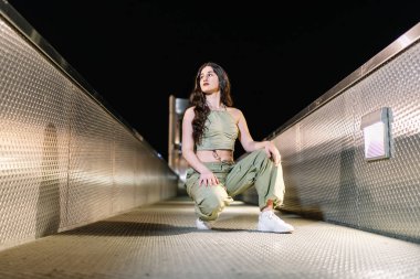 Sokak dansçısı poz veriyor ve metal bir köprüye bakıyor, şehir ışıklarıyla aydınlatılıyor, şehir gece hayatının özünü yakalıyor.