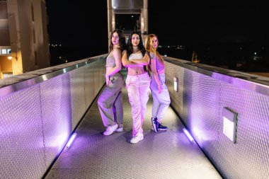 Üç genç bayan sokak dansçısı, mor ışıklarla aydınlatılmış bir köprüde poz veriyor.