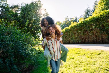 Genç bir adam, neşeli bir parkta kız arkadaşına sırtına biniyor, hem gülüyor, hem de güneşli bir günün sıcaklığını emiyor.