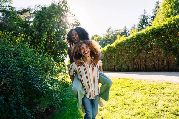 Genç bir adam, neşeli bir parkta kız arkadaşına sırtına biniyor, hem gülüyor, hem de güneşli bir günün sıcaklığını emiyor.