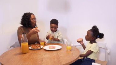 Anne ve çocuklar birlikte kahvaltı yapıyor, çörek yiyor, hamur işi yiyor ve meyve suyu içiyor.