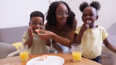 Bakıcı anne iki küçük çocuğa besleyici sabah yemeği servis ediyor, mutfakta yemek yerken sevgi ve beraberlik anlarını paylaşıyor.