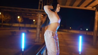 Enerjik bayan dansçı, geceleri mavi neon ışıklarıyla aydınlatılan bir köprünün altında hip hop hareketleri sergiliyor.
