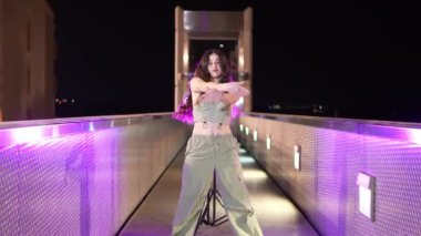 Aydınlatılmış köprü üzerinde sokak dansı yapan enerjik bayan dansçı, dinamik hareket ve sanatsal ifade yoluyla canlı şehir gece hayatını sergiliyor.