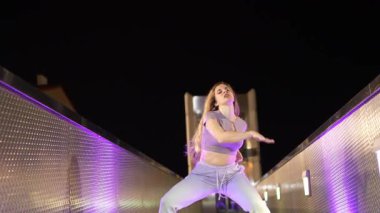 Gece vakti şehir köprüsünde dinamik sokak dansı koreografisi yapan genç bayan dansçı, modern hareket ve sanatsal ifadeyi enerji ve kendine güvenle sergiliyor.