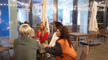 Dostlar bir masada toplanır, sıcak bir gece ortamında içki, yemek ve canlı sohbetin tadını çıkarırlar.