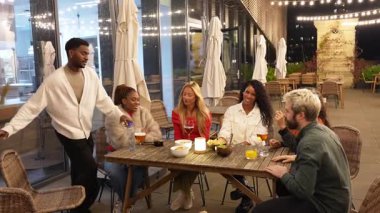 Bir grup farklı arkadaş canlı çatı katı barında buluşuyor, gülüşüyor, sarılıyor ve içkilerini paylaşıyor.