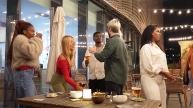 Şehir merkezindeki çatı katı barında canlı sohbetler ve içkileri paylaşan arkadaşlar. Şehir ışıkları da arka planda.