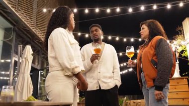 Genç yetişkin arkadaşlar gece vakti sosyal toplantının keyfini çıkarıyorlar, içkilerini ve sohbetlerini açık terasta sıcak ip ışıkları altında paylaşıyorlar.