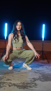 Kendine güvenen genç bir kadın geceleri çatıda çeşitli hip hop dansları yapıyor. Mavi neon ışıklarla aydınlatılıyor.