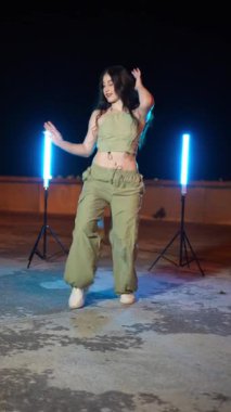 Genç bayan dansçı şehir çatılarında hareketli hip hop hareketleri sergiliyor. Gece vakti sokak performansı sırasında parlak neon ışıklarıyla aydınlatılıyor.