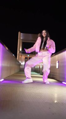 Mor ışıklarla aydınlatılmış bir köprüde çeşitli hip hop hareketleri sergileyen enerjik bayan dansçı.