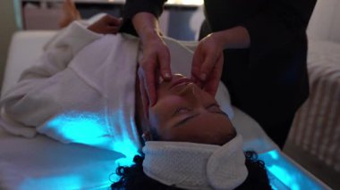 Profesyonel güzellik uzmanının masaj masasında uzanan genç bir kadına uyguladığı yavaş çekim yüz masajı.