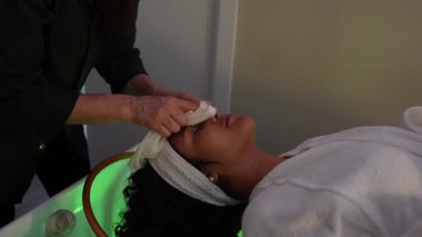 Bir güzellik uzmanının, bir sağlık merkezinde masaj yatağında uzanan genç bir kadının yüzünü nazikçe temizlerken çekilmiş yavaş çekim videosu.