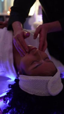 Yavaş çekim yüz masajı, profesyonel güzellik uzmanı tarafından güzellik kliniğinde, renkli ışıklarla genç bir kadına yapılır.