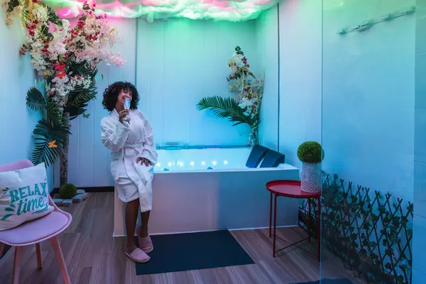 Bornoz giyen, şampanya içen ve modern bir güzellik merkezinde rahatlayan genç bir kadın.