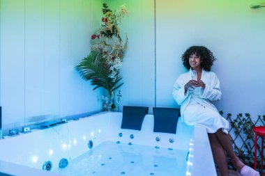 Güzellik uzmanı modern bir masaj küvetinde rahatlatıcı bir anın tadını çıkarıyor, spa tedavisi yaptıktan sonra ferahlatıcı bir içecek tutuyor.