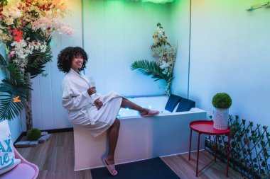 Genç bir kadın renkli bir kaplıcada bir bardak köpüklü şarapla bir hidromasaj küvetinin kenarında oturarak rahatlamanın tadını çıkarıyor.