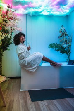 Müşteri, renkli ışıkları ve çiçekli süslemeleri olan bir kaplıcadaki duyusal yoksunluk tankının kenarında otururken bir bardak şampanyanın tadını çıkarıyor.