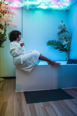 Modern güzellik merkezinde aydınlanmış küvetin kenarında otururken bornoz giyen ve şampanya içen genç bir kadın.