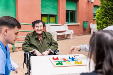 Hemşire ve arkadaşlarıyla parcheesi oynamaktan hoşlanan yaşlı bir kadın, bir huzurevinde sosyal etkileşimi ve bilişsel uyarımı teşvik ediyor.