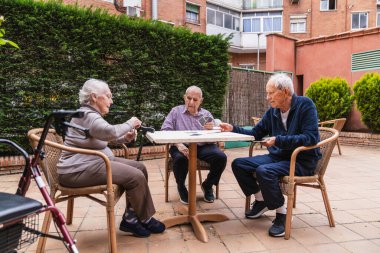 Yaşlı bireyler bir huzurevinin bahçesindeki açık bir masada oyun oynarlar, sosyal etkileşimi ve rahatlamayı teşvik ederler.