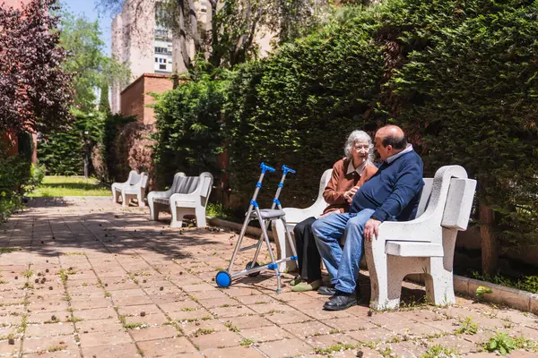Huzurlu bir huzurevinin bahçesindeki bankta sohbetten zevk alan aile yakınlarda yürüyüp destek ve hareketlilik sağlıyor.