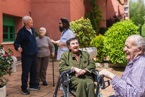 Yaşlı insanlar güneşli bir günün tadını çıkarıyorlar, sohbet ediyorlar ve gülüyorlar. Huzurevinin bahçesinde dost canlısı bir hemşire yardım ediyor.