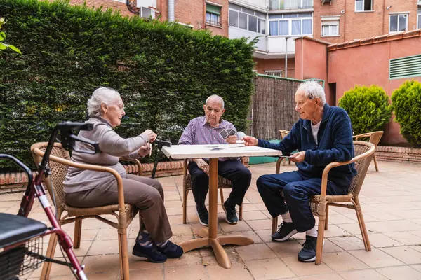 Yaşlı bireyler bir huzurevinin bahçesindeki açık bir masada oyun oynarlar, sosyal etkileşimi ve rahatlamayı teşvik ederler.
