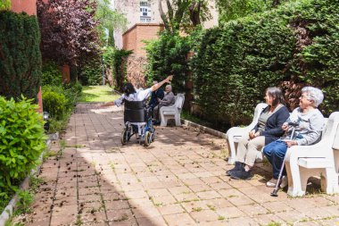 Hemşire yaşlı bir kadını tekerlekli sandalyeye itiyor, güneşli bir günün tadını çıkarırken, bir huzurevinin canlı bahçesiyle çevrili.