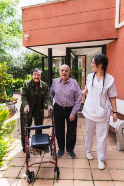 Hemşire, bir huzurevinin bahçesinde gezinirken, destek ve hareketlilik için bir aylak kullanarak neşeli yaşlı bir çifte yardımcı oluyor.