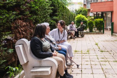 Hemşire gülümsüyor ve bir huzurevinin bahçesindeki bankta oturan iki yaşlı kadınla konuşuyor.