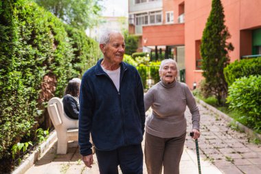 Mutlu yaşlı çift birlikte bir huzurevinin bahçesinde yürüyüş yapıyor, açık hava ve arkadaşlığı kucaklıyor.